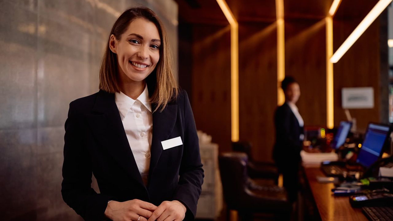 Smiling hotel receptionist standing at a softly lit front desk.