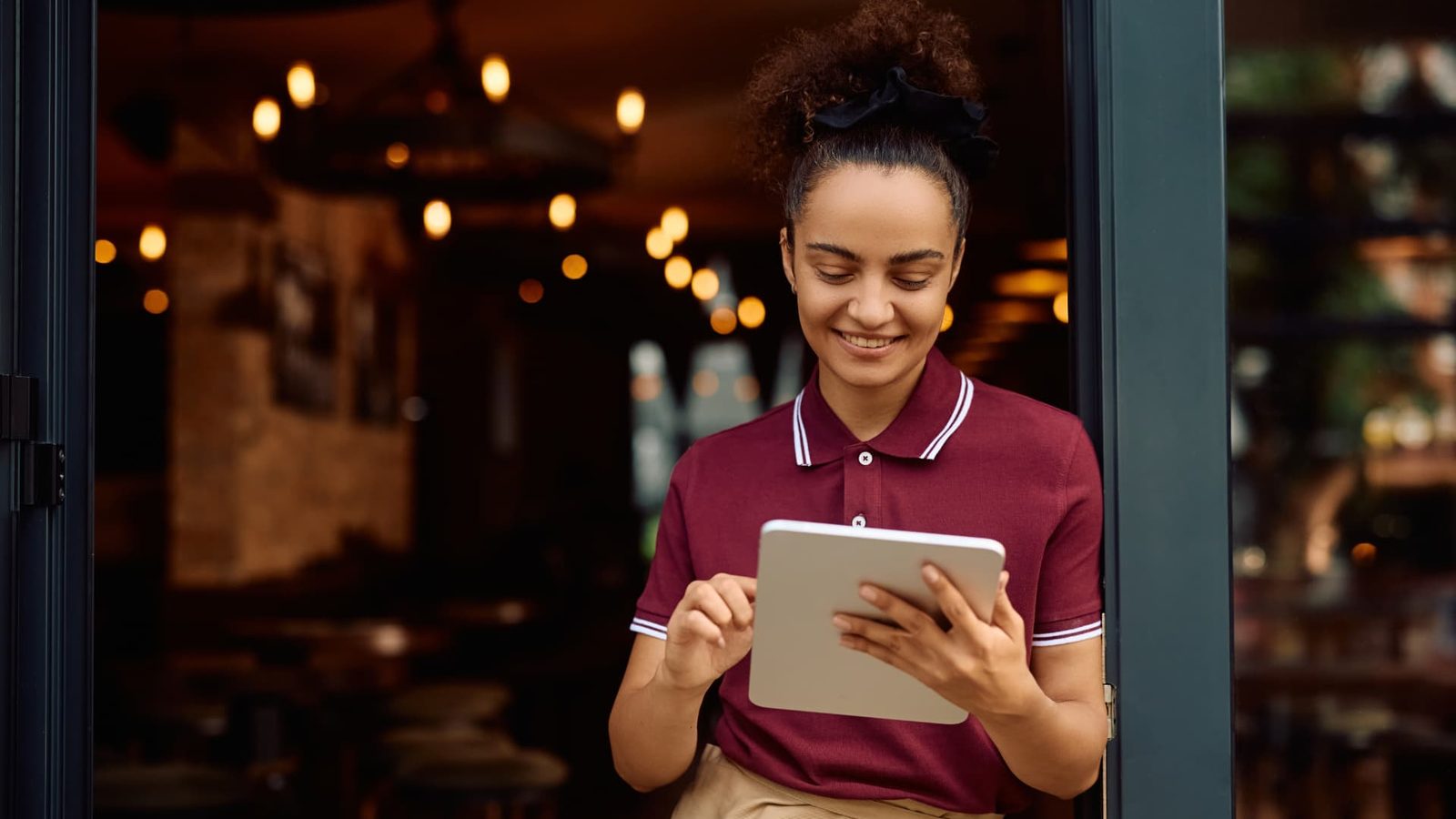 Smiling hospitality server in a burgundy polo shirt using a tablet in the doorway of a warmly lit restaurant.