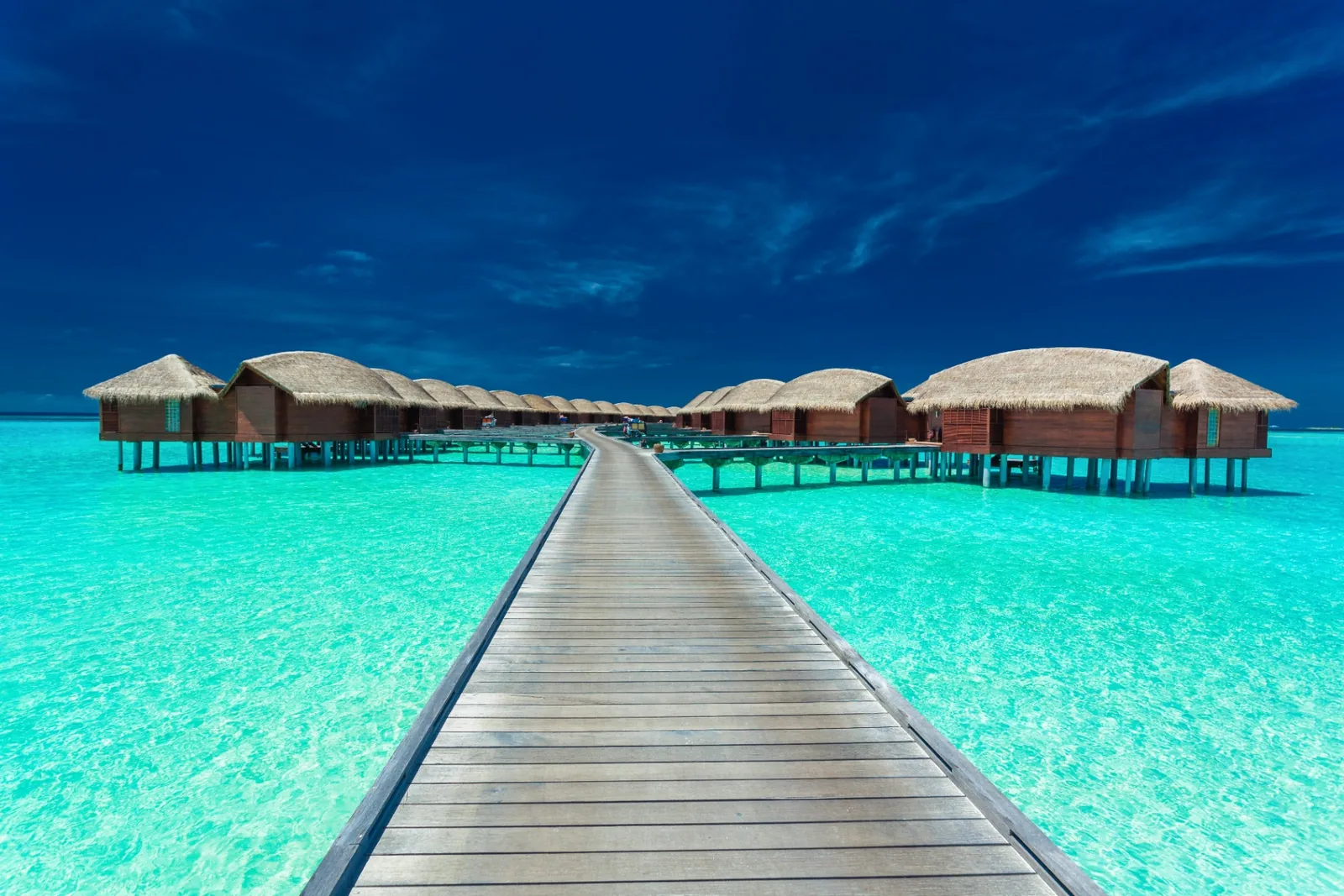 Wooden boardwalk leading to thatched-roof overwater villas above a turquoise Maldivian lagoon