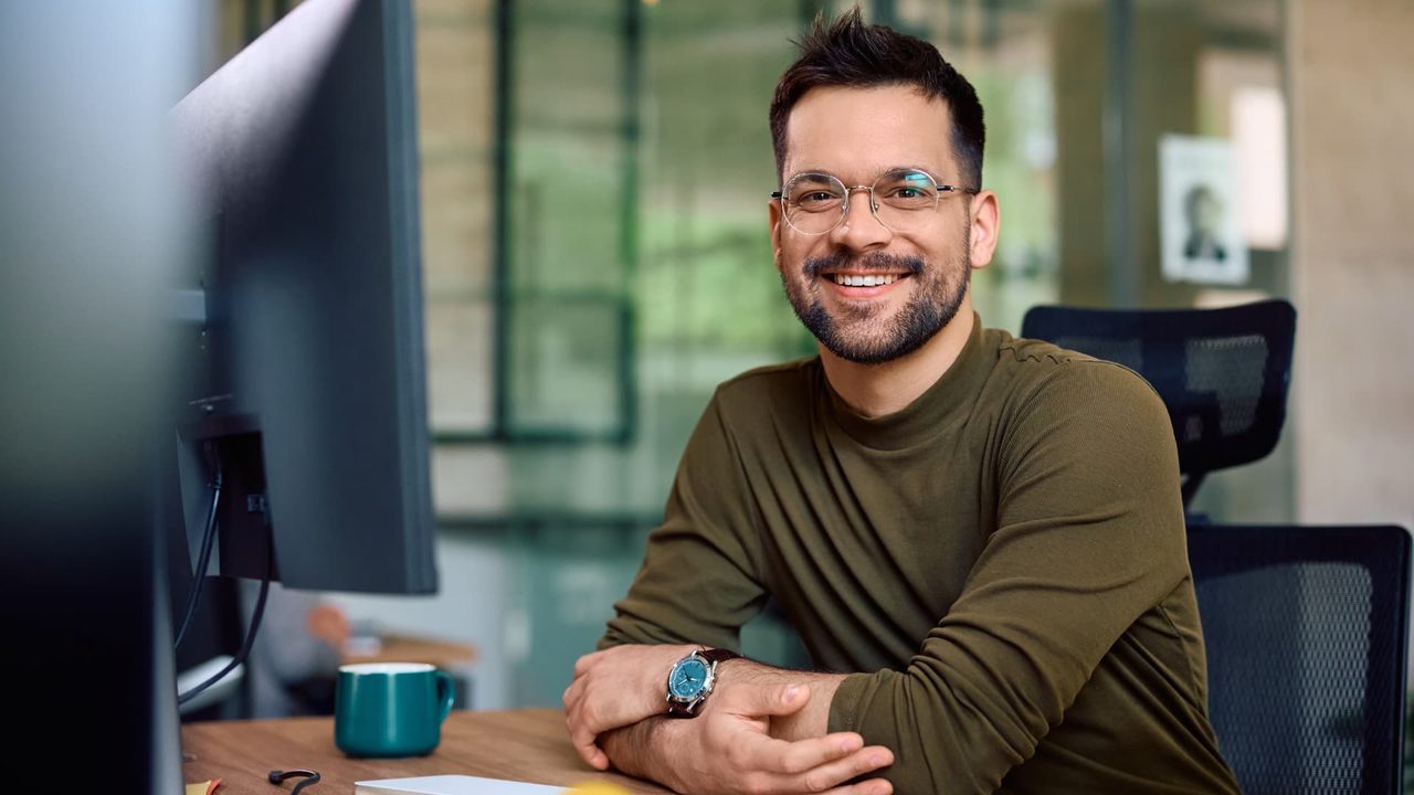 Smiling developer in glasses sitting at a desk with a monitor and keyboard in a bright modern office.