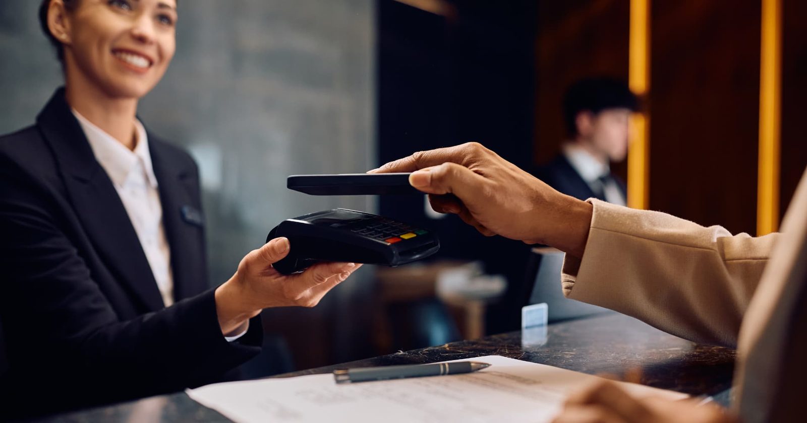 Hotel guest paying contactlessly with a phone on a payment terminal held by a receptionist at the front desk